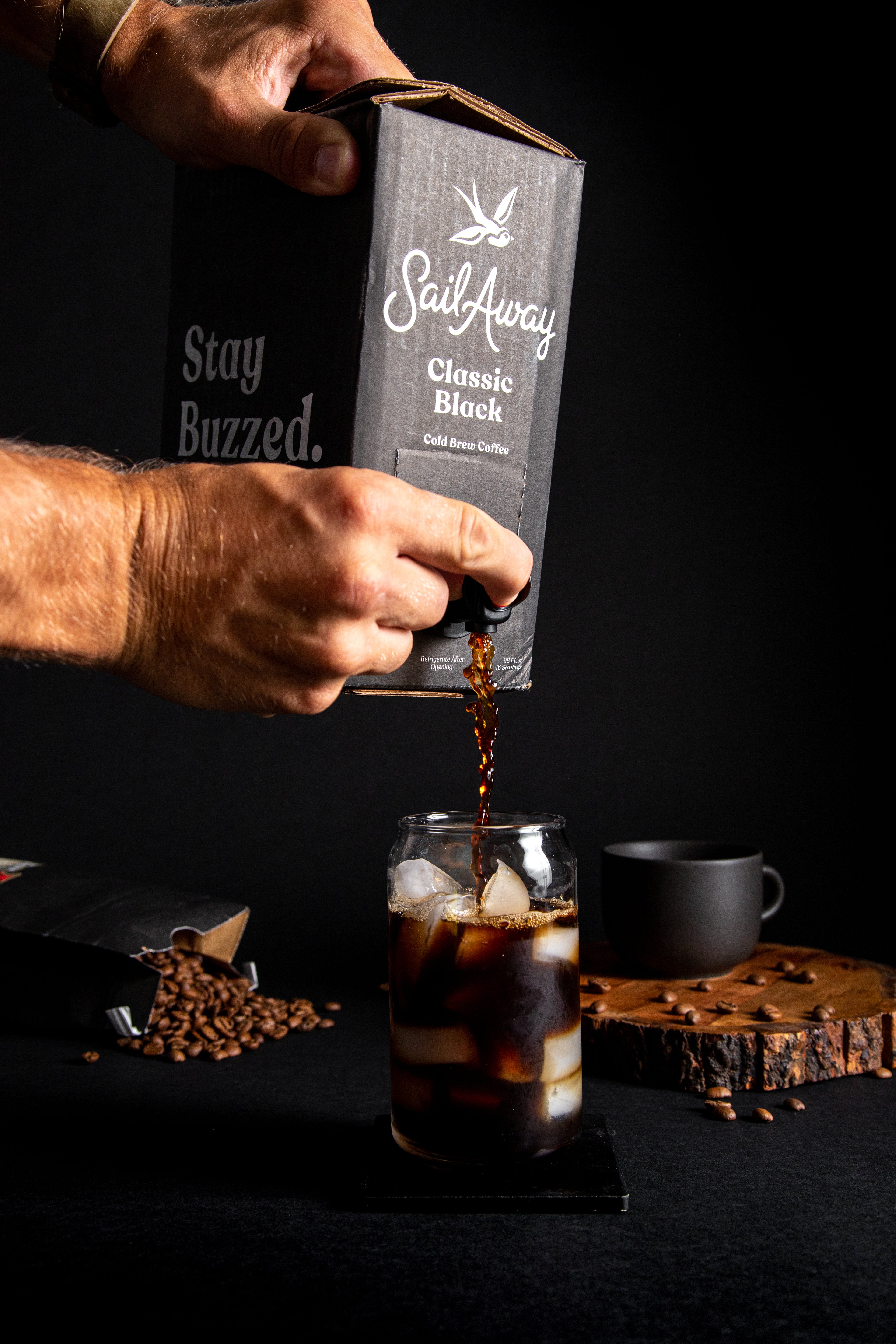 Hand pouring coffee from a 'Sail Away' coffee bag into a glass with ice, on a dark background.